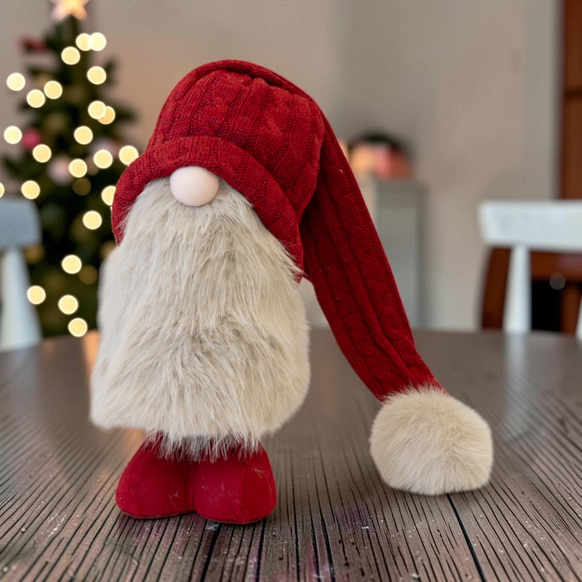 Large red standing Christmas gonk with cable-knit hat and faux-fur beard on a table by a lit Christmas tree