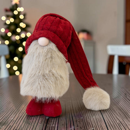 Large red standing Christmas gonk with cable-knit hat and faux-fur beard on a table by a lit Christmas tree