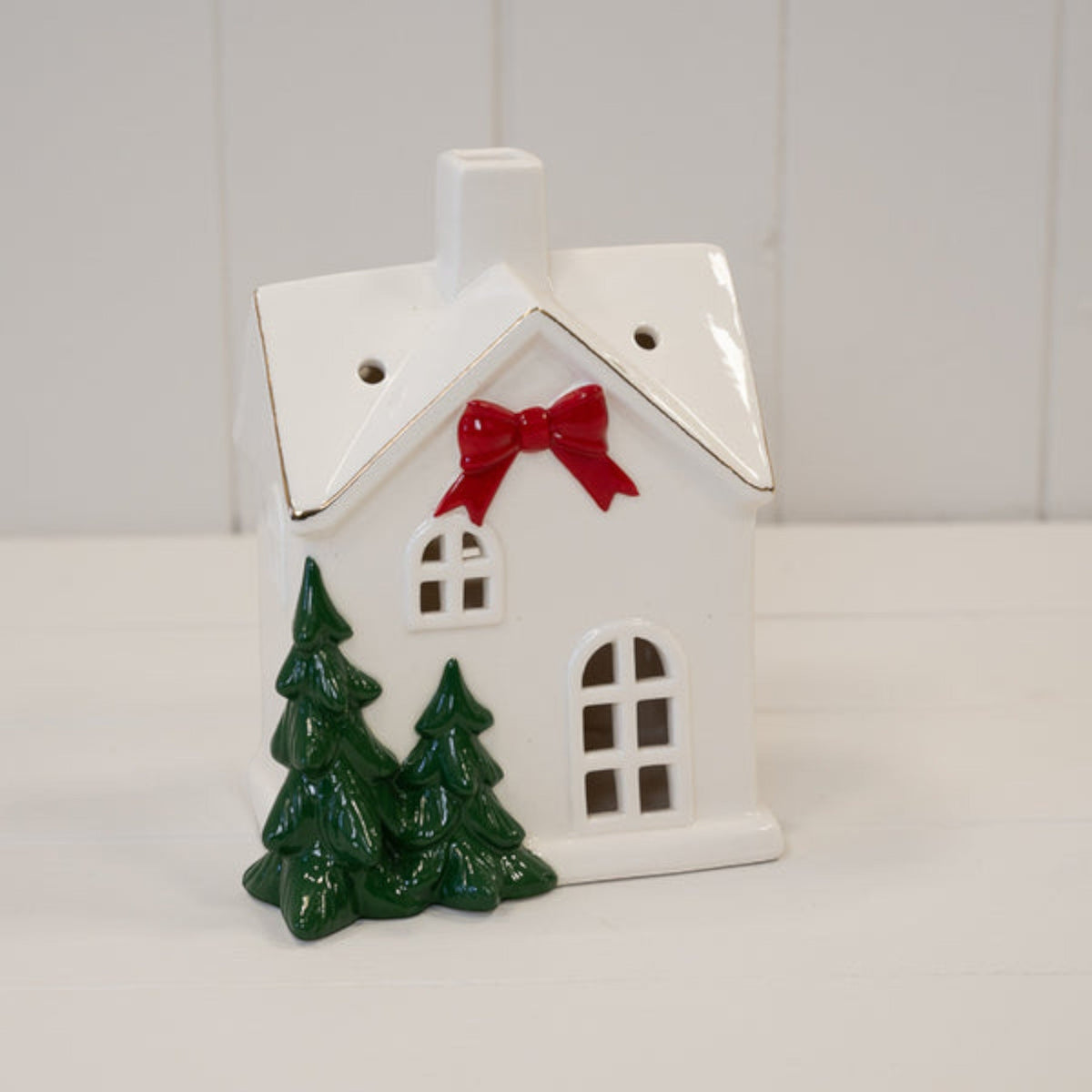 Large white ceramic Christmas house with red bow and fir trees on a white shelf