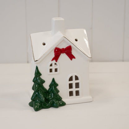 Large white ceramic Christmas house with red bow and fir trees on a white shelf