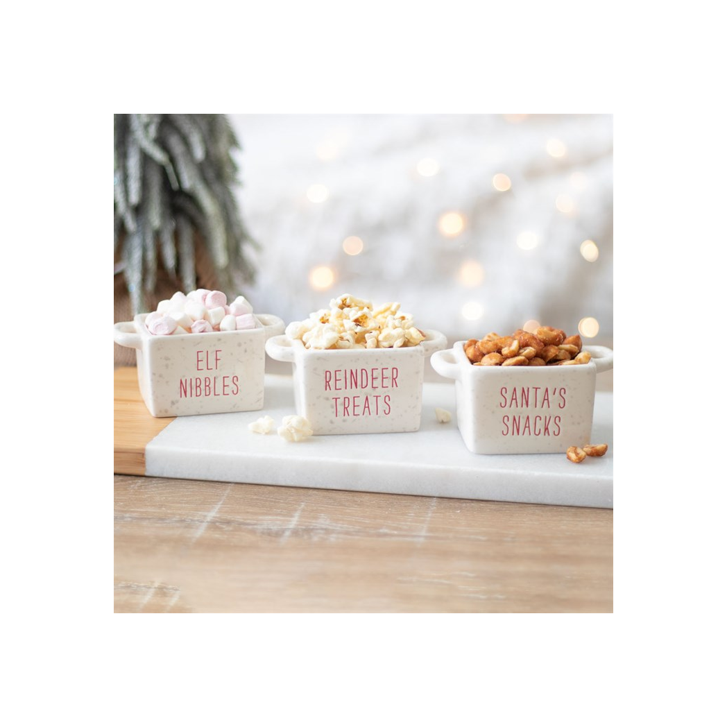 Three Christmas snack bowls filled with marshmallows, popcorn and pretzels on a marble board