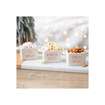 Three Christmas snack bowls filled with marshmallows, popcorn and pretzels on a marble board