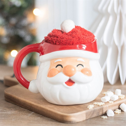 Santa face mug topped with red fluffy socks and a pompom on a wooden board with marshmallows